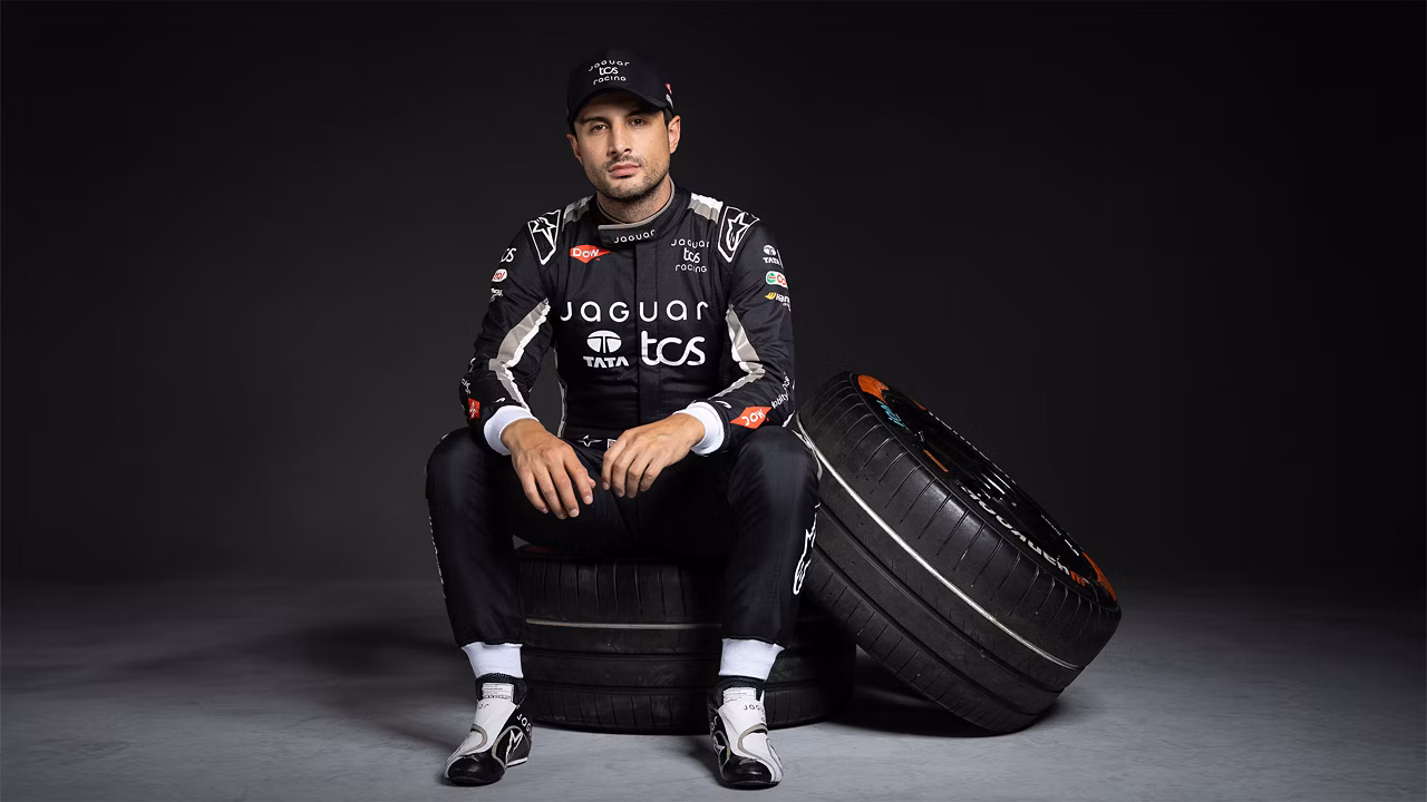 A racing driver in a Jaguar TCS Suit sits on a tire with another tire beside him.