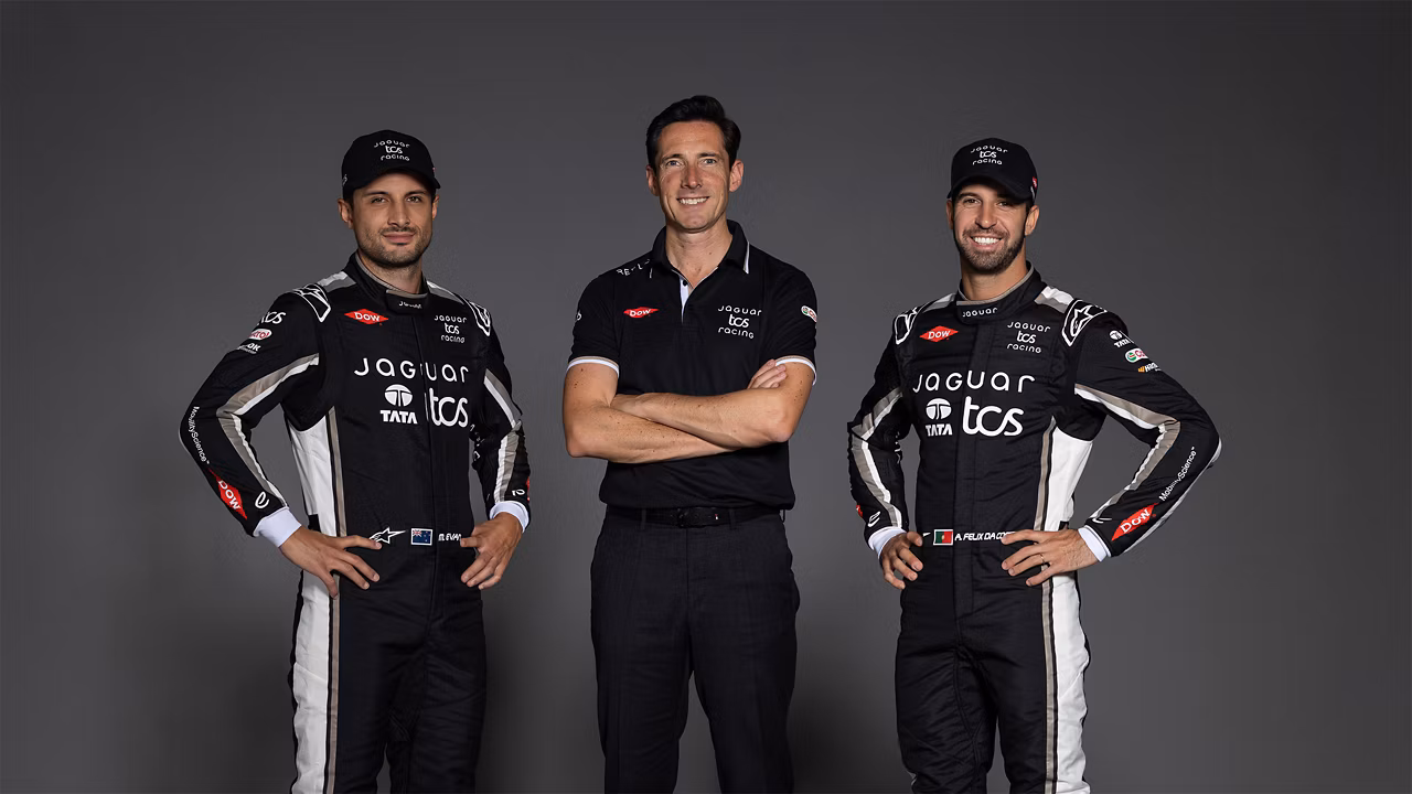 Three men pose in a studio, two in a Jaguar TCS racing suit stand on either side of one in a black team shirt.