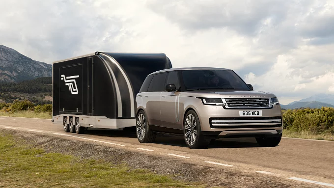 Range Rover towing a large, aerodynamic black enclosed trailer along a country road against a mountain backdrop.